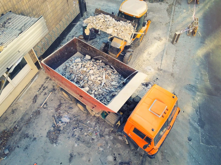 Chantier de démolition extérieur avec pelleteuse orange et bleue détruisant un mur en briques rouges