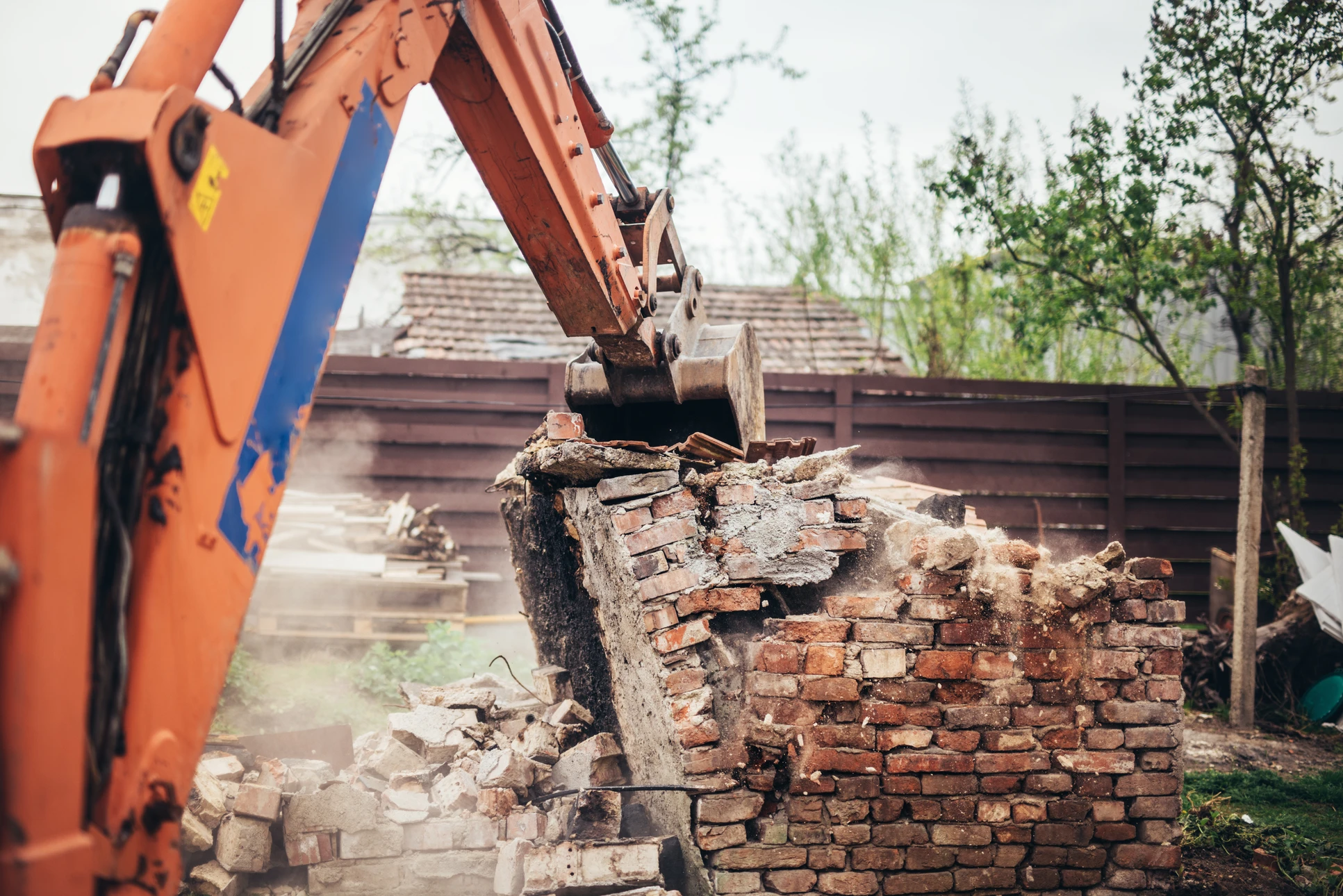 Chantier de démolition extérieur avec pelleteuse orange et bleue détruisant un mur en briques rouges